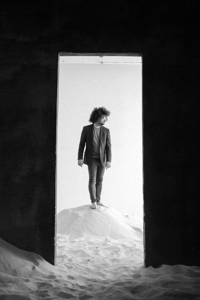 Michael S.U. Hudson, Creator, standing in a doorway on sand, black and white